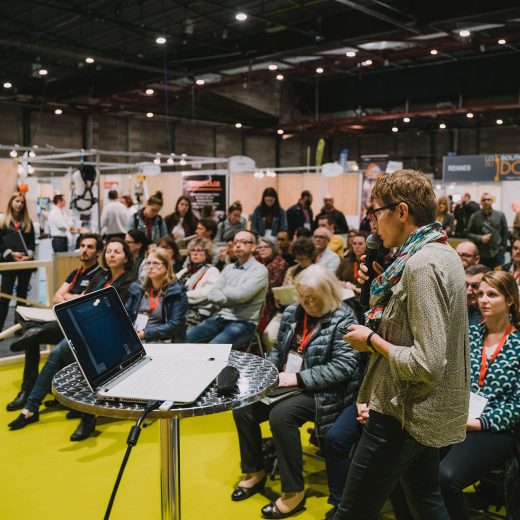 Une femme parle dans un micro face à un écran hors champ, des personnes l’écoutent assises derrière elle Une femme parle dans un micro face à un écran hors champ, des personnes l'écoutent assises derrière elle